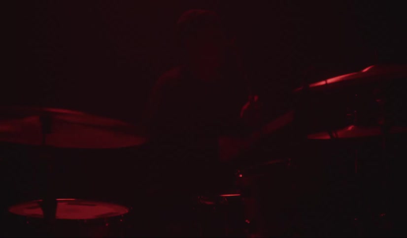 Male musician wearing a toque plays the drums and cymbals with drumsticks as lights flash on him