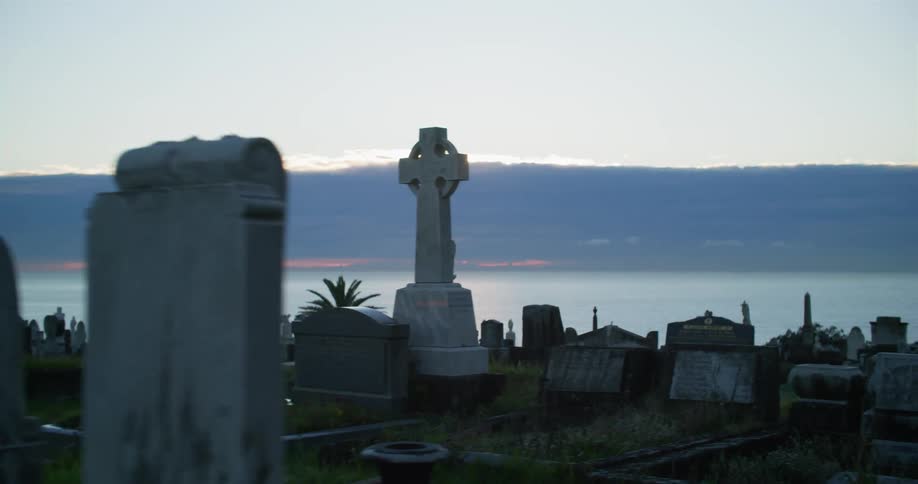 Clouds obscure red tinted sky on horizon of ocean in background behind cemetery