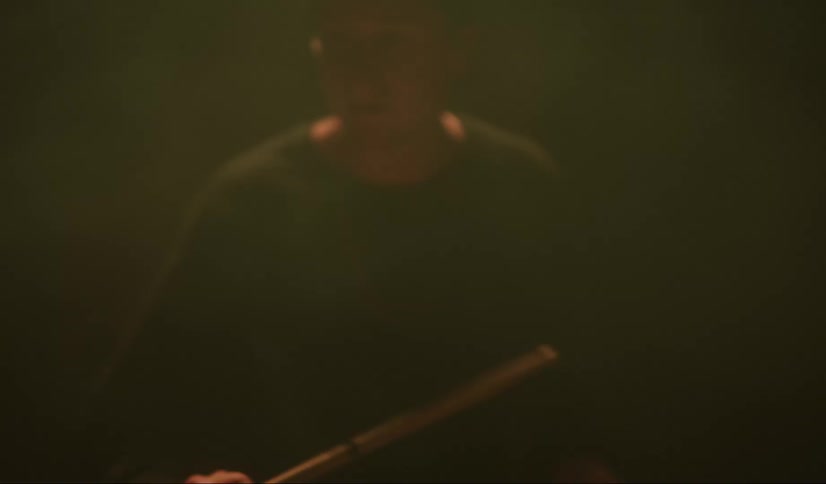 A man in a room sitting playing the drums with drumsticks with a wall in the background