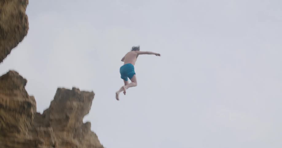 Two men standing on a cliff and one of the men jumping into water in a lagoon with the sky and clouds in the background