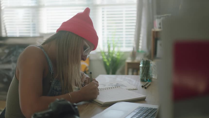 Woman wearing glasses and a pink toque writes in a notebook as a laptop and camera sit nearby