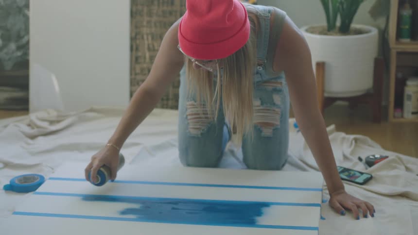 An artist sprays blue paint onto a taped canvas on a tarp on the floor