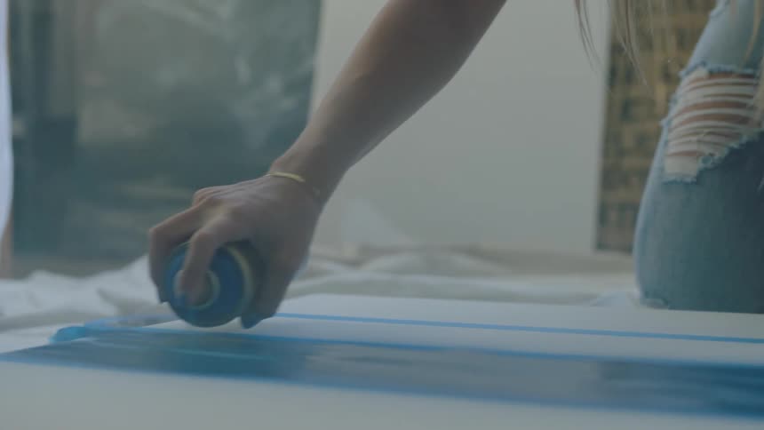 Blonde female artist kneels on spread fabric spray painting onto canvas