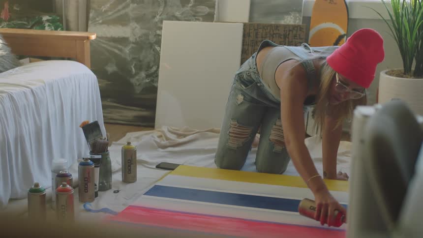 A woman spray paints on a canvas on a tarp on the floor