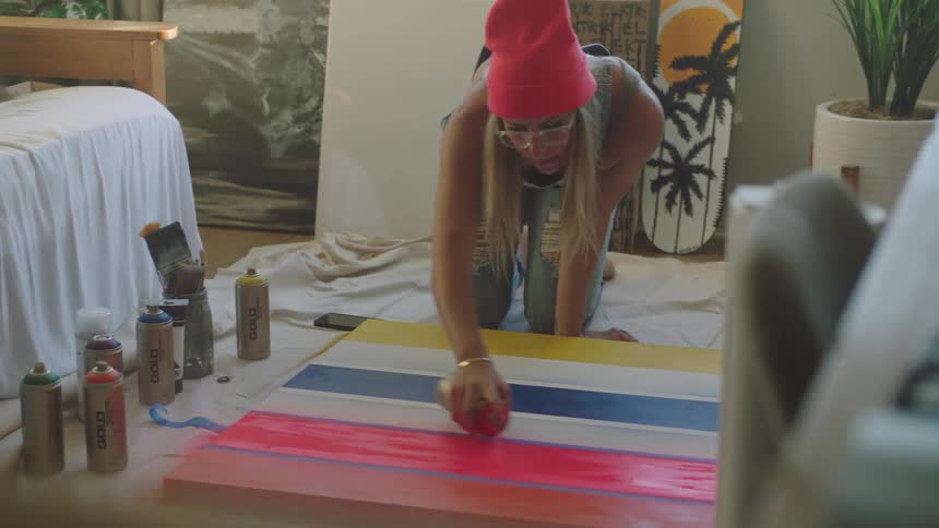 Woman wearing glasses and a pink toque spray paints stripes onto a canvas while kneeling on the floor