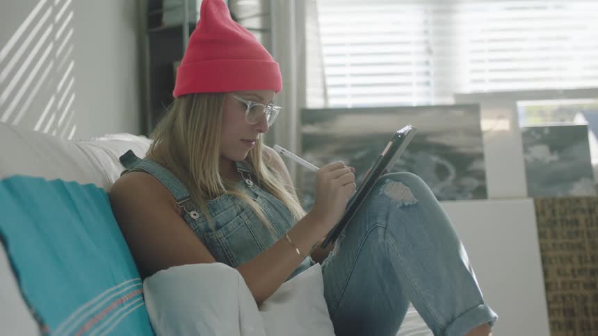 A woman walks across a room and sits on a sofa before beginning to write on a computer tablet with a stylus