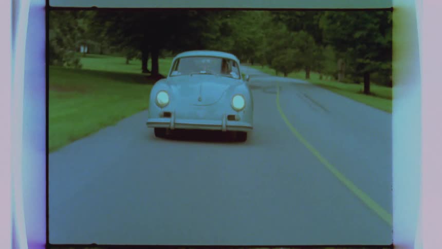 An older blue car drives down a wooded road