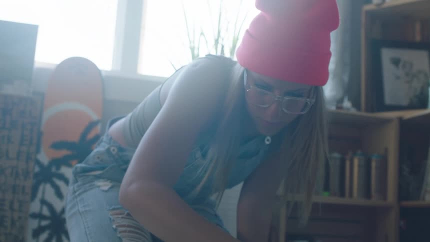 Woman wearing glasses and a toque sprays paint onto a canvas that is on the floor