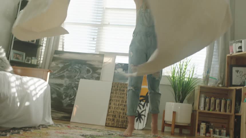 A woman with a hat and glasses spreading a sheet on the floor with a window in the background