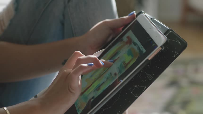 A woman operates art software on a computer tablet with her stylus while sitting on a sofa indoors during the day