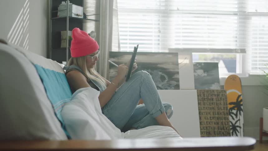 A woman with a hat and glasses sitting on a couch using a tablet with a stylus