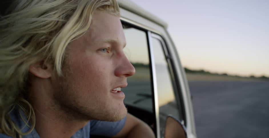 A man leans out the window of a moving car at sunset