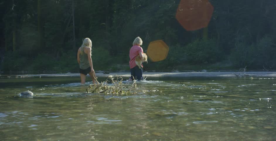 Two women walk across a stream and one carries a dog
