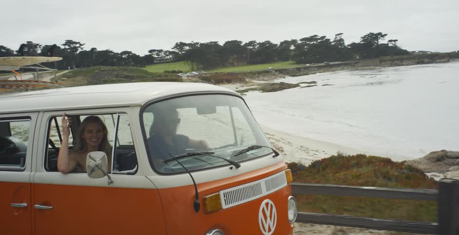 A man and woman are driving in a v.w. bus along a coastal road in daylight