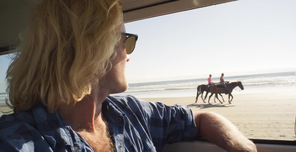 Blond man in a vehicle looks out the window at people riding horses on the beach