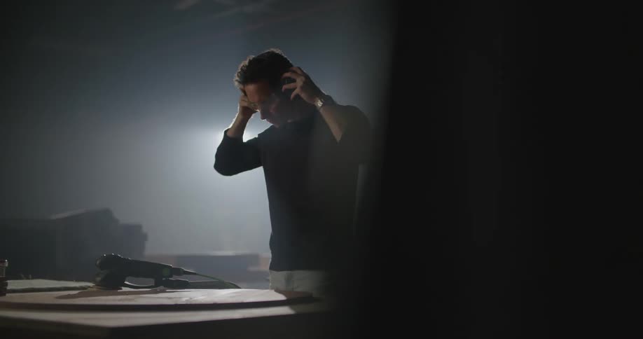 A man in a workshop with goggles and earmuffs picking up an electric sander with a light in the background