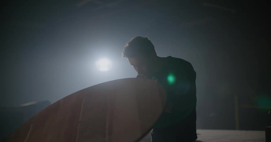 Man blowing the sawdust from a piece of wood he is working on inside a dimly lit warehouse