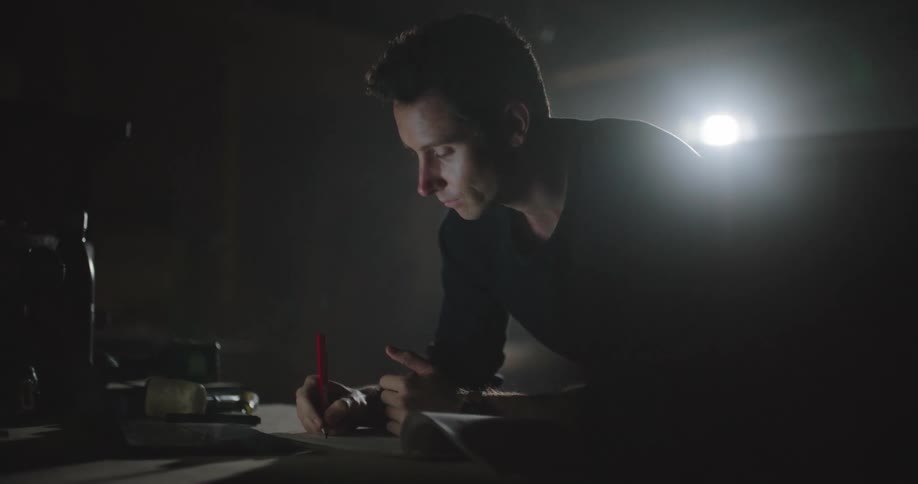 Young man working on a laptop and writing over a desk inside a dimly lit room