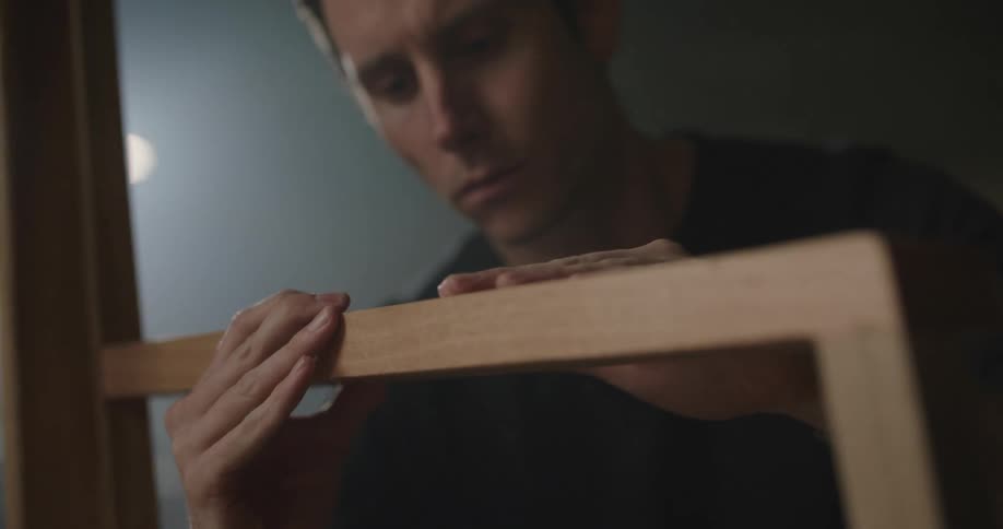 Young man checks the finished surface of a chair inside a dimly lit warehouse