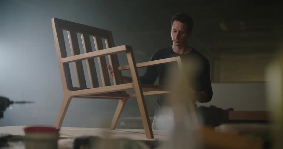 A carpenter places a wooden chair on a table and polishes the arms with a towel