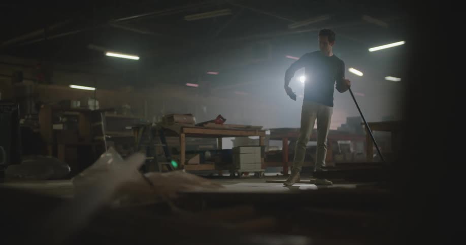 A man is sweeping the floor of a workshop and then pauses to take a phone call