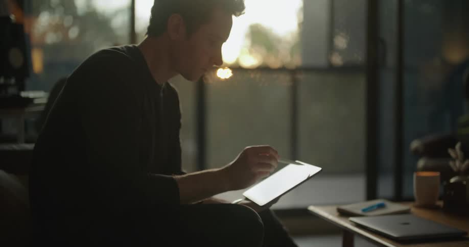 Man sits on couch as sun shines through balcony doors sketching on tablet