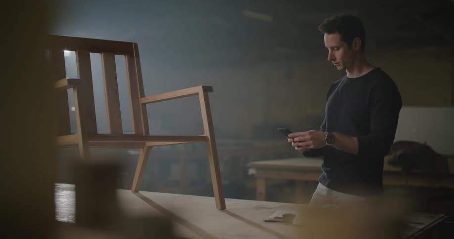 Man stands in workshop near wooden chair with clipboard inspecting it before taking pictures using a smartphone