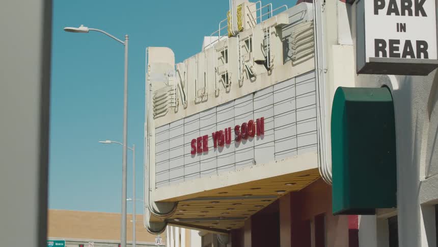 A movie theater with a sign in front with buildings the sky and clouds in the background