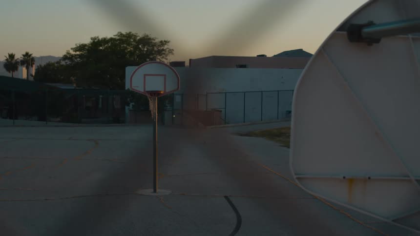 Chain link fence encloses lot with basketball court with buildings in background