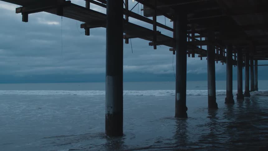 Ocean waters form eddies around pier supports as waves roll onto beach under overcast sky