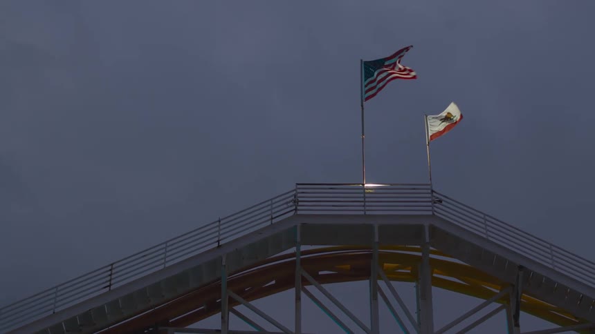 A roller coaster track with the american flag and the california flag blowing in the wind with the sky and clouds in the background