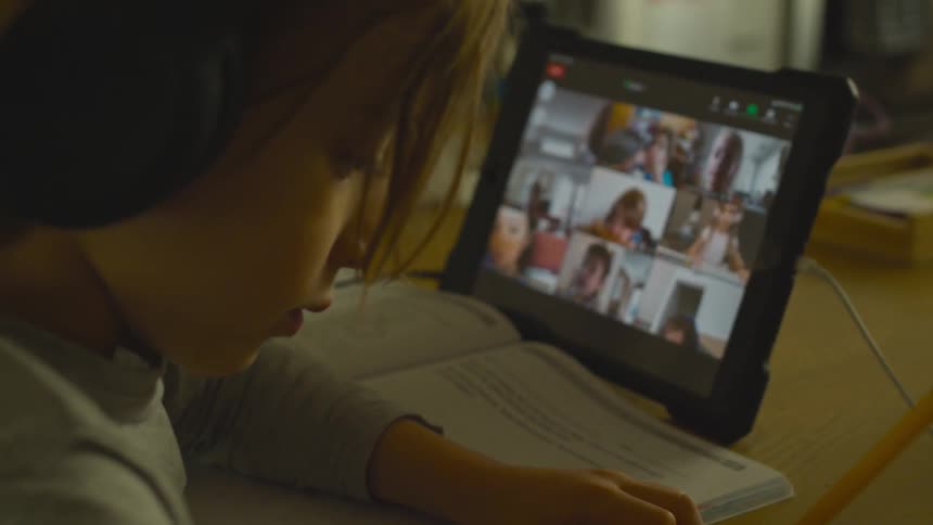 A little boy with headphones sitting at a table holding a pencil with a book and a tablet next to him