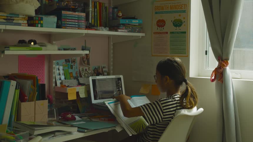 Teenaged girl holds a book and holds up four fingers while looking at a monitor on the desk