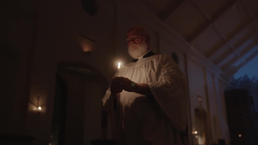 An older religious figure walking down and lit only by a candle that he's carrying