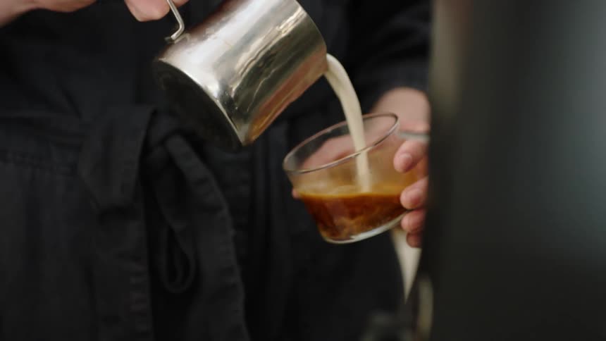A person pours cream from a small kettle into a class coffee cup