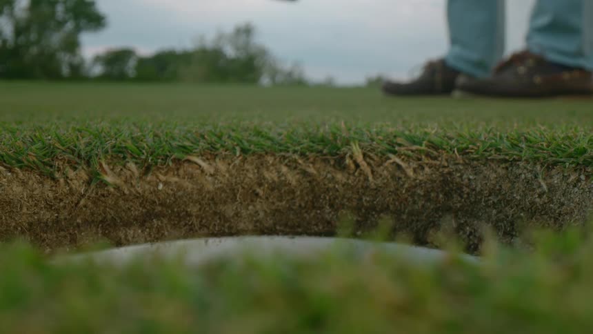 Man with jeans and brown shoes hits golf ball into hole on the grass