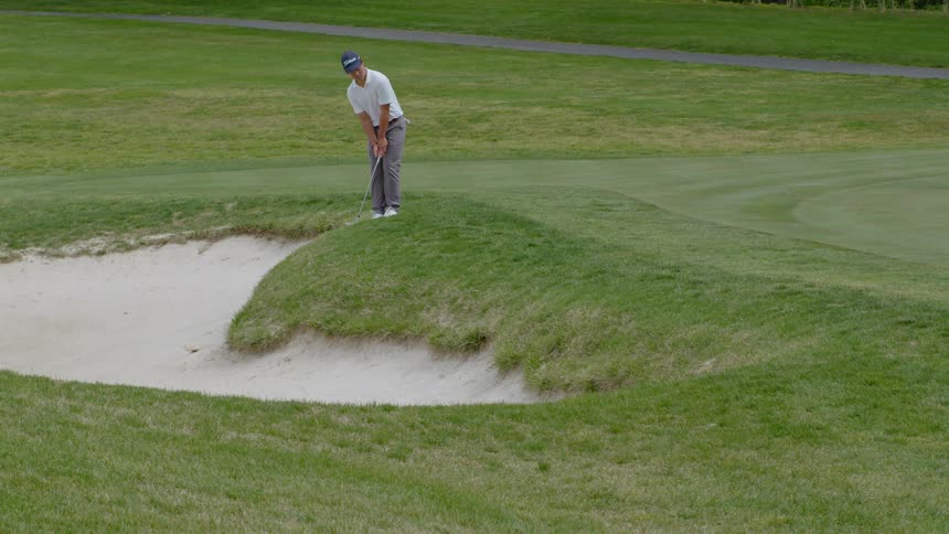 A man strikes a golf ball with his club while standing on the edge of a sand bunker on a golf course during the day
