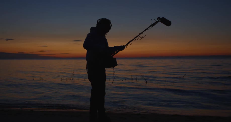 A man operates a boom mic on a dark beach at sunset