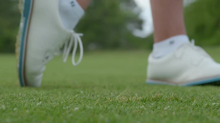A person with shoes and socks swinging a golf club at a golf ball with trees the sky and clouds in the background