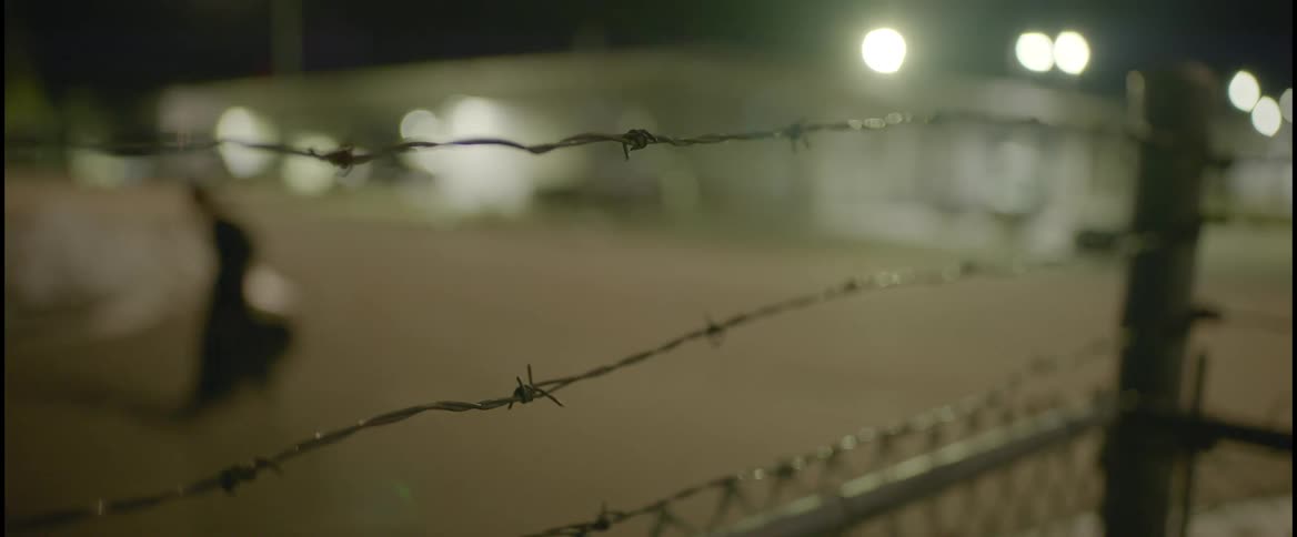 A fence with barbed wire with a person walking buildings and lights in the background