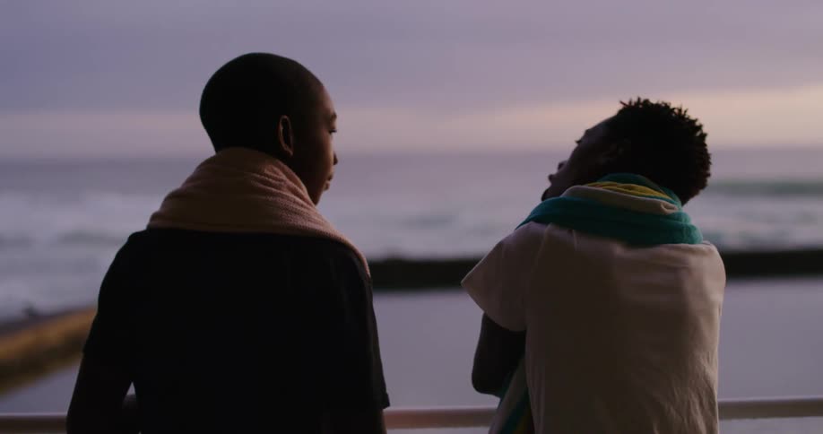 One teenage boy demonstrates breathing and swimming stroke to another while they stand outdoors near enclosed pond with ocean waves crashing onto shore