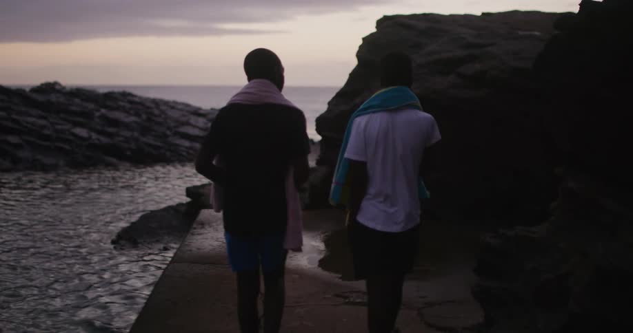 Two boys with towels walking near the coast with the ocean sky and clouds in the background