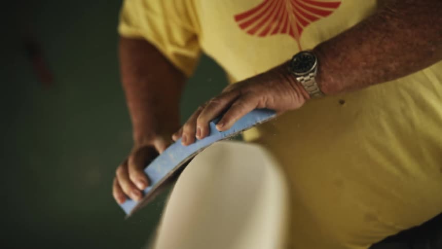 A man uses a large sanding pad to sand an unfinished surfboard