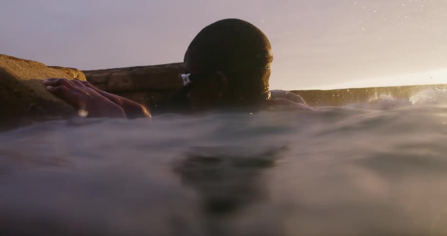 Black male swimmer wearing goggles clings to a rock in the ocean