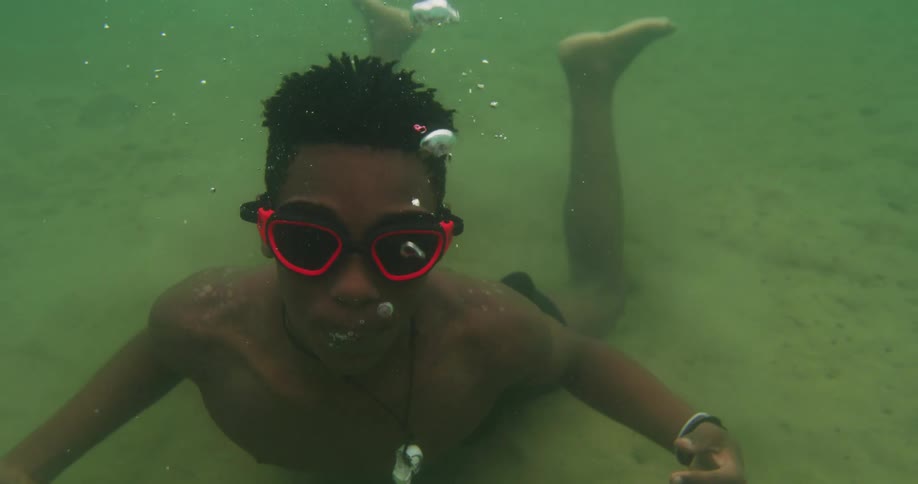 Man wearing goggles swims below the surface of the ocean and exhales bubbles