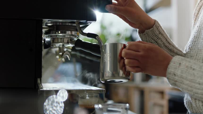 Steam billows from an espresso machine as a barista uses a steam wand to froth milk