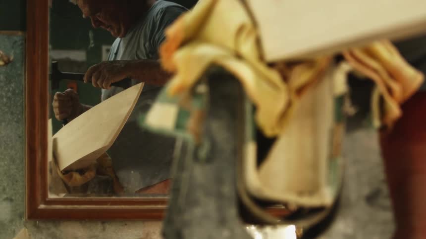 A craftsman holding a mallet working on a clamped piece of wood is reflected in the mirror on his workshop wall