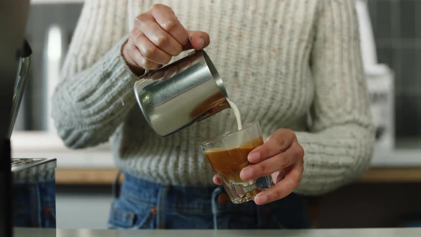 Barista wearing a sweater and jeans creates latte art with small metal pitcher and glass