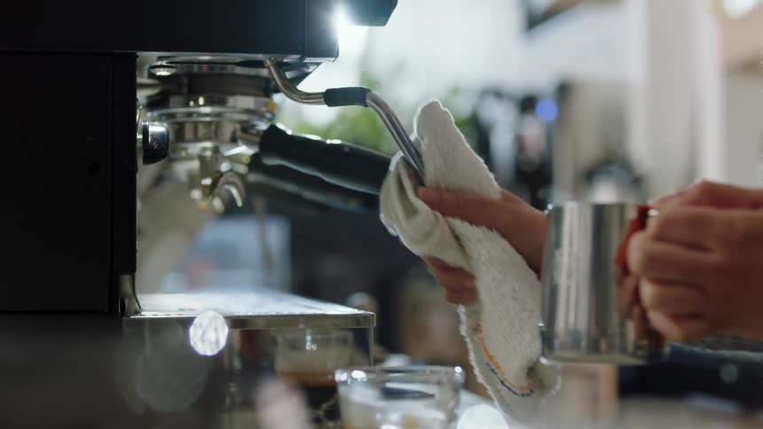 A person in a room with a sweater using a cappuccino machine and wiping it with a towel