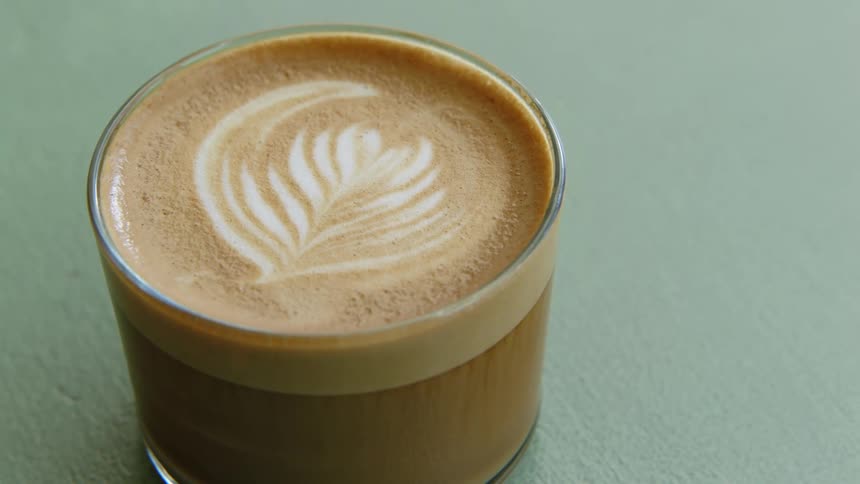 Latte art topped beverage sits in low glass on textured pale green surface